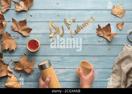 Hands holding insulated metal flask and bamboo cup. Zero waste tea in Fall. Flat lay on faded light wood planks with scattered Autumn leaves. Stock Photo