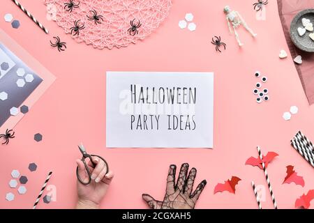 Halloween party ideas text on white page held in hand. Flat lay with scissors and decorations on pink paper background. Paper drink straws, bats, suga Stock Photo