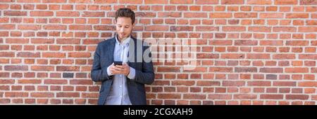 Businessman sms texting phone app in city street on brick wall background. Business man holding smartphone in smart casual wear standing. Urban young Stock Photo