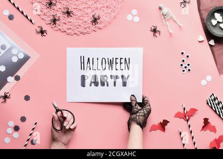 Halloween party text on white page held in hand. Flat lay with scissors and decorations on pink paper background. Paper drink straws, bats, sugar hear Stock Photo