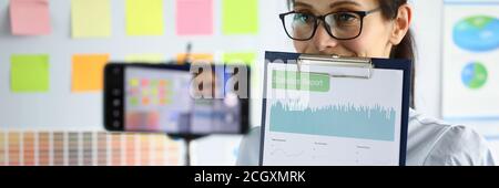 Woman in office holds charts in her hand and demonstrates them through a smartphone Stock Photo