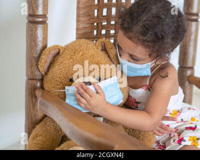 A girl in a white dress is putting a mask on her teddy bear Stock Photo