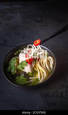 Spicy asian soup taken by chopsticks. Chicken bouillon and meat, red chilli pepper, cilantro, scallions, japanese noodles in black soup bowl on black Stock Photo