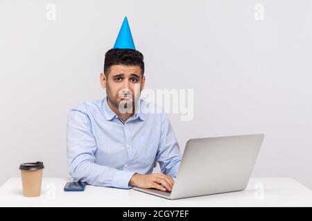 Unhappy employee guy with funny party cone on head sitting in office workplace, using laptop and looking depressed, upset about canceled weekend. indo Stock Photo