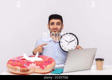 Time to rest! Upset man showing clock, looking frustrated tired at camera, sitting in office workplace with rubber ring passport and paper airplane on Stock Photo