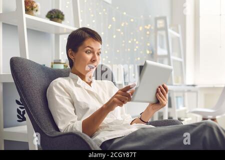 Overjoyed adult woman reading exciting news on tablet sitting in comfortable armchair at home Stock Photo