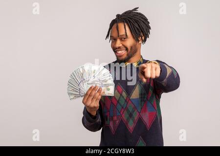 Happy satisfied african guy with dreadlocks holding lot of money in hand and pointing finger at you, inviting to participate in lottery. Indoor studio Stock Photo