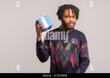 Disappointed curious african guy with dreadlocks trying to guess what is inside gift box, sudden present, delivery service. Indoor studio shot isolate Stock Photo