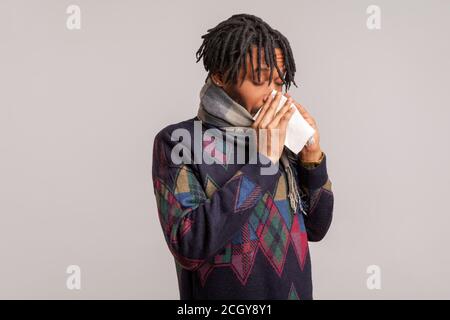 Side view african guy with dreadlocks blowing his nose into napkin, suffering cold and runny nose, seasonal epidemic. Indoor studio shot isolated on g