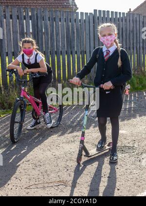 Two ten year old schoolgirls in school uniform one on a bike and the other on a scooter both wearing face masks. Stock Photo