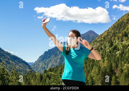 Stock photo of a 30's woman with face mask taking a selfie with phone while enjoying the mountains and a beautiful landscapes on a sunny day Stock Photo