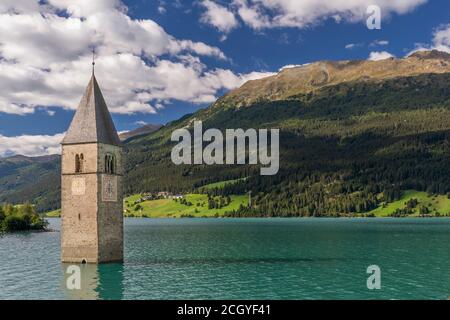 The ancient submerged bell tower of Lake Resia in Val Venosta, South Tyrol, Italy, emerges from the water against a beautiful blue sky with white clou Stock Photo