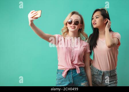 Multi ethnic female models having fun, fooling around, taking selfie at studio over blue background. Stock Photo