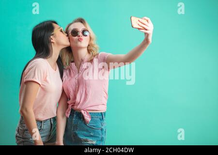 Thai young woman kissing his caucasian blonde girlfriend in sunglasses with duck lips while shooting selfie isolated on blue background. Stock Photo