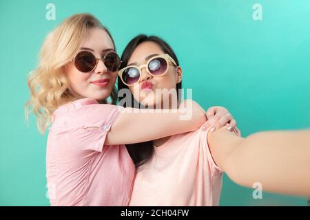 Portrait of pretty charming girls in casual outfits, wearing sunglasses shooting selfie , making duck faces, using smart phone isolated on studio blue Stock Photo