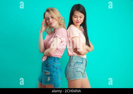 Two multiracial women being in conflict, standing back to back over blue background. Indoor studio shot of angry and dissapointed females in bad mood. Stock Photo
