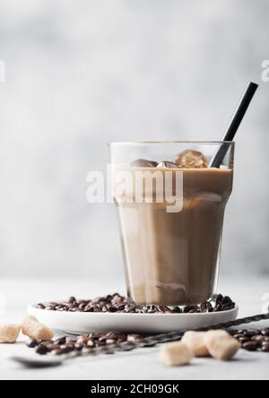 Iced cold coffee with milk and raw beans and spoon with coffee beans in white plate on light table background with cane sugar. Stock Photo