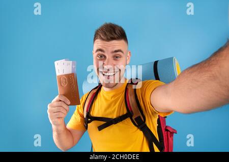 Happy young man with backpack, passport and plane tickets taking selfie on blue background Stock Photo