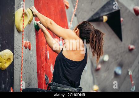 Extreme sport, stress relief, bouldering, people and healthy lifestyle concept. Young sporty muscular woman climbing up on top of rock wall in gym, si Stock Photo