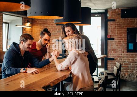 Joyful young couple in burger pub loughing and testing their strengths sitting at table doing arm wrestling with happy smiles. Their friends cheering Stock Photo