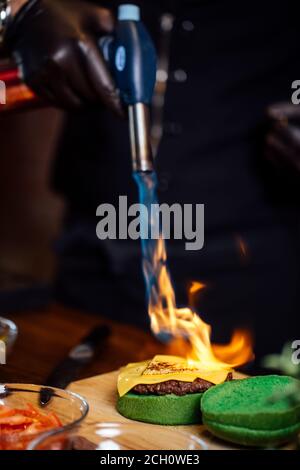 Chef making beef burgers. Melting cheese on burger bun with culinary burner for burgers at restaurant kitchen. Stock Photo