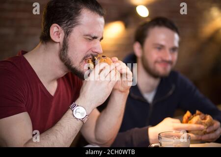 Two happy male friends sitting, talking and having lunch with burgers and beer at cozy cafe interior. One emotional guy making a good bite of tasty bu Stock Photo