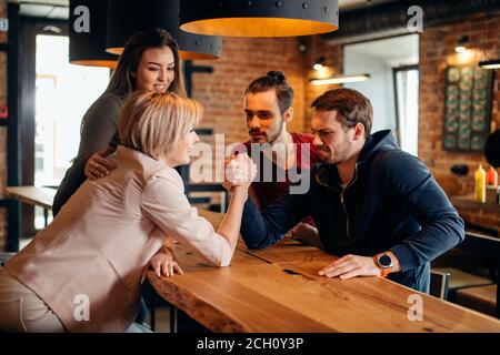 Cheerful group of students friends couple having fun arm wrestling each other in fashionable pub. Stock Photo