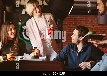 Young woman with cocktail suggesting a toast. Group of young people enjoying time in popular bar. Youth and leisure, Pastime and Party concept. Stock Photo