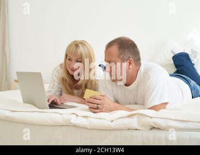 Mature family couple sitting at bed in modern home, looking at laptop screen. Happy middle aged man holding credit card and female making purchases online Stock Photo