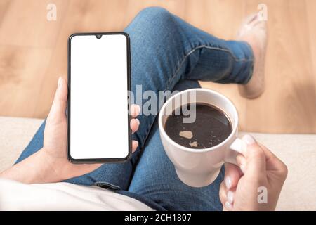 mockup cellphone. Mockup image of woman's hands holding mobile phone with blank screen on thigh and coffee cup in home Stock Photo