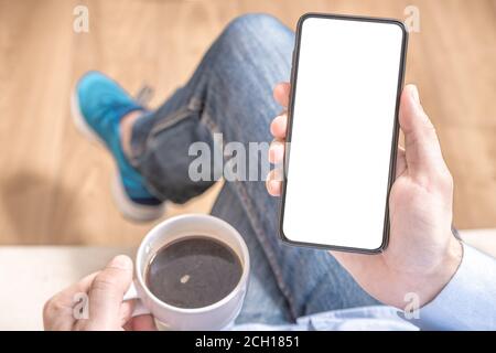 Mockup cellphone. Mockup image of men's hand holding mobile phone with blank screen on thigh and coffee cup. Stock Photo
