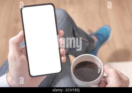 Top view mockup image of a man holding a black mobile phone with blank white desktop screen with coffee cup. Cell phone mockup. Stock Photo