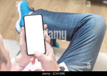 mockup cellphone. Man sitting on a sofa and holding a blank screen mobile phone. Man wearing jeans relaxing at home, lying on comfortable sofa, readin Stock Photo
