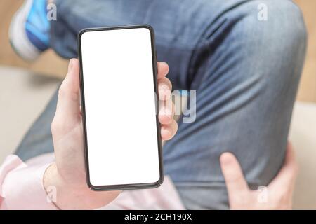 Mockup image blank white screen cell phone. Man hand holding texting using mobile on sofa at home. Empty space for advertise text. Young man in jeans Stock Photo