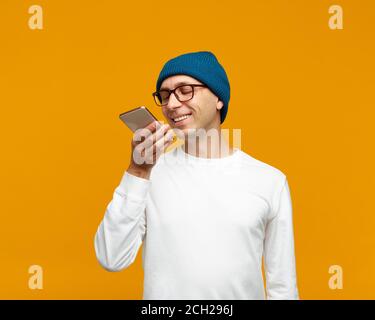 Portrait of positive young man recording audio message, speaking to microphone of mobile phone. Male isolated on bright yellow colored background. White jumper and blue hat. Stock Photo