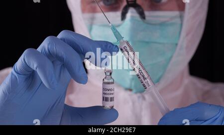Coronavirus vaccine. Man in white safety uniform, mask and glasses holding coronavirus vaccine ampoule and syringe. COVID-19 pandemic cure concept