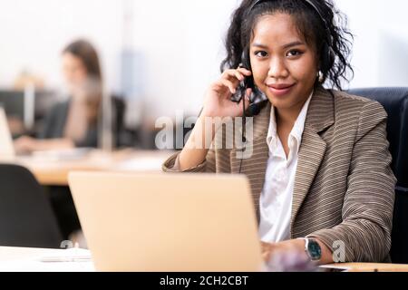 Young adult friendly confidence operator Mixed race of African and asian woman with headsets working in a call center with her colleague in background Stock Photo