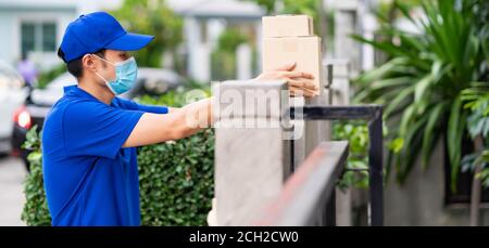 Panoramic side view asian deliver man with face mask in blue shirt handling packages and put in on the fence post as contactless shopping delivery. Th Stock Photo