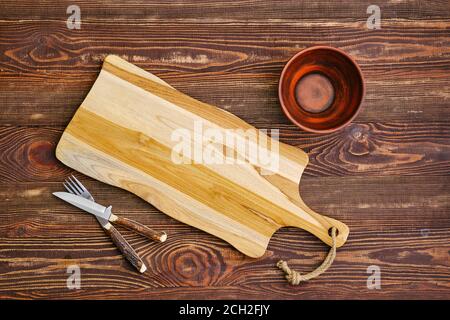 Top view of empty cutting wooden board with clay bowl and fork with knife on the table Stock Photo