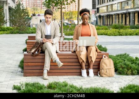Two young busy colleagues of various ethnicities using mobile gadgets outdoors Stock Photo