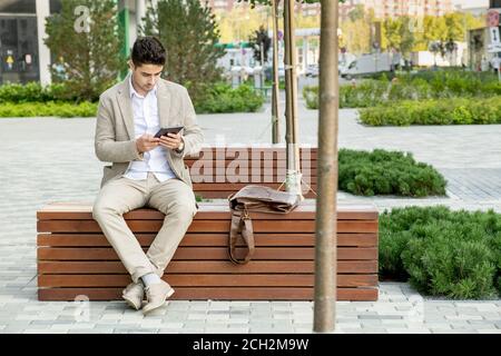 Young contemporary businessman in formalwear networking on wooden bench in park Stock Photo