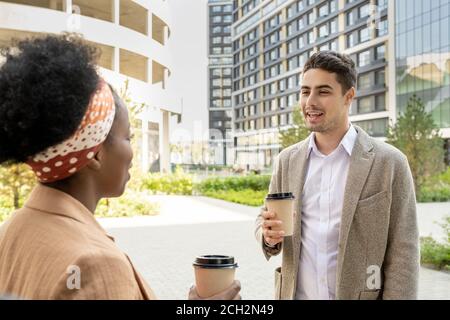 Happy young businessman with glass of coffee talking to African female colleague Stock Photo