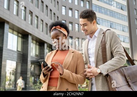 Young smiling businessman looking at screen of smartphone in hands of colleague Stock Photo