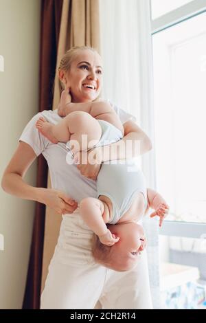 Happy mother playing with her baby son at home holding infant upside down. Family having fun