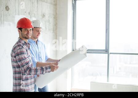 handsome bearded builder is looking at the camera while his colleague is looking at the blueprint of the new office. panorama window on the background Stock Photo