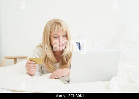 Middle age female on online shopping. Portrait of Mature beautiful blond woman is holding credit card and using laptop computer laying on bed at home Stock Photo