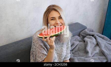Young attractive woman bites a piece of watermelon. Woman at home in a cozy interior looking at camera and smiling Stock Photo