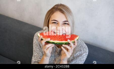 Young attractive woman bites a piece of watermelon. Woman at home in a cozy interior looking at camera and smiling Stock Photo