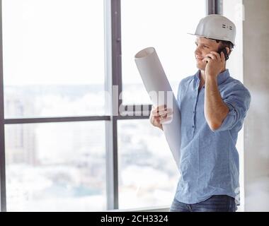 business conversation on the phone.smiling builder in casual outfit and hardhat is using his cell phone at work. side view photo. panorama window on t Stock Photo