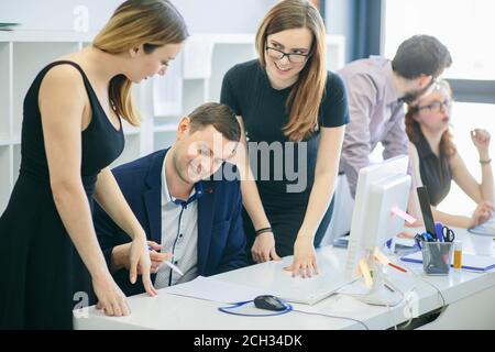 female employees are standing over Caucasian chief sitting at the table indoors. sly workers are cheating their boss with contract Stock Photo
